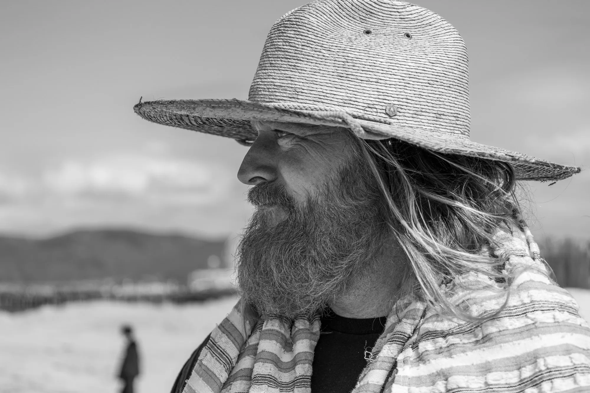 A bearded man in a wide-brimmed hat looks toward the sea in a black-and-white portrait.