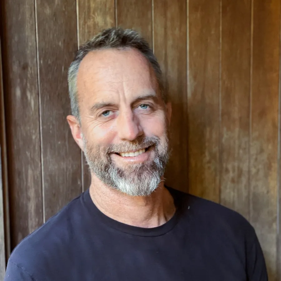 A bearded man smiles in front of a weathered wooden door.