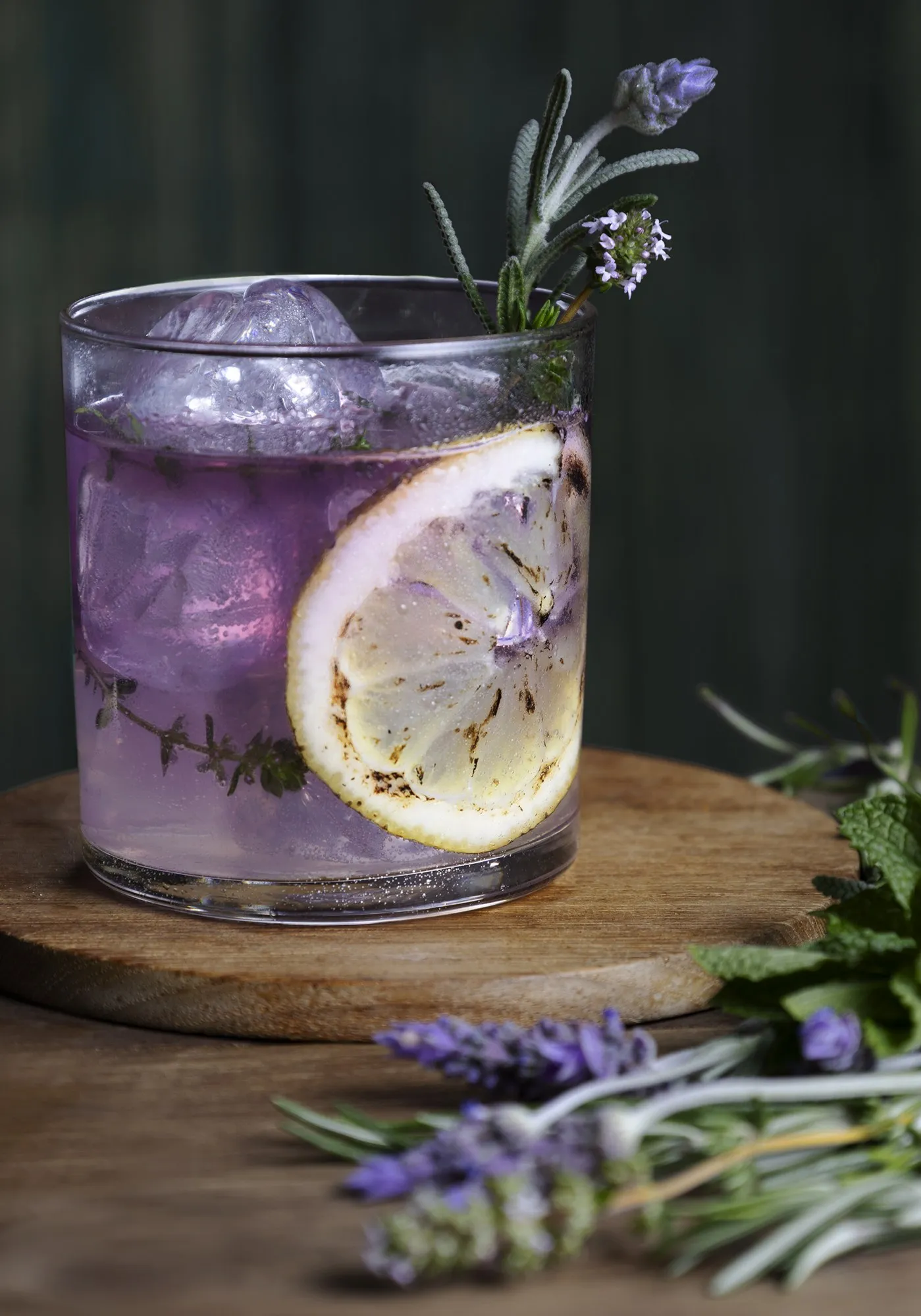 A lavender cocktail with lemon and herbs sits on a dark wooden table.