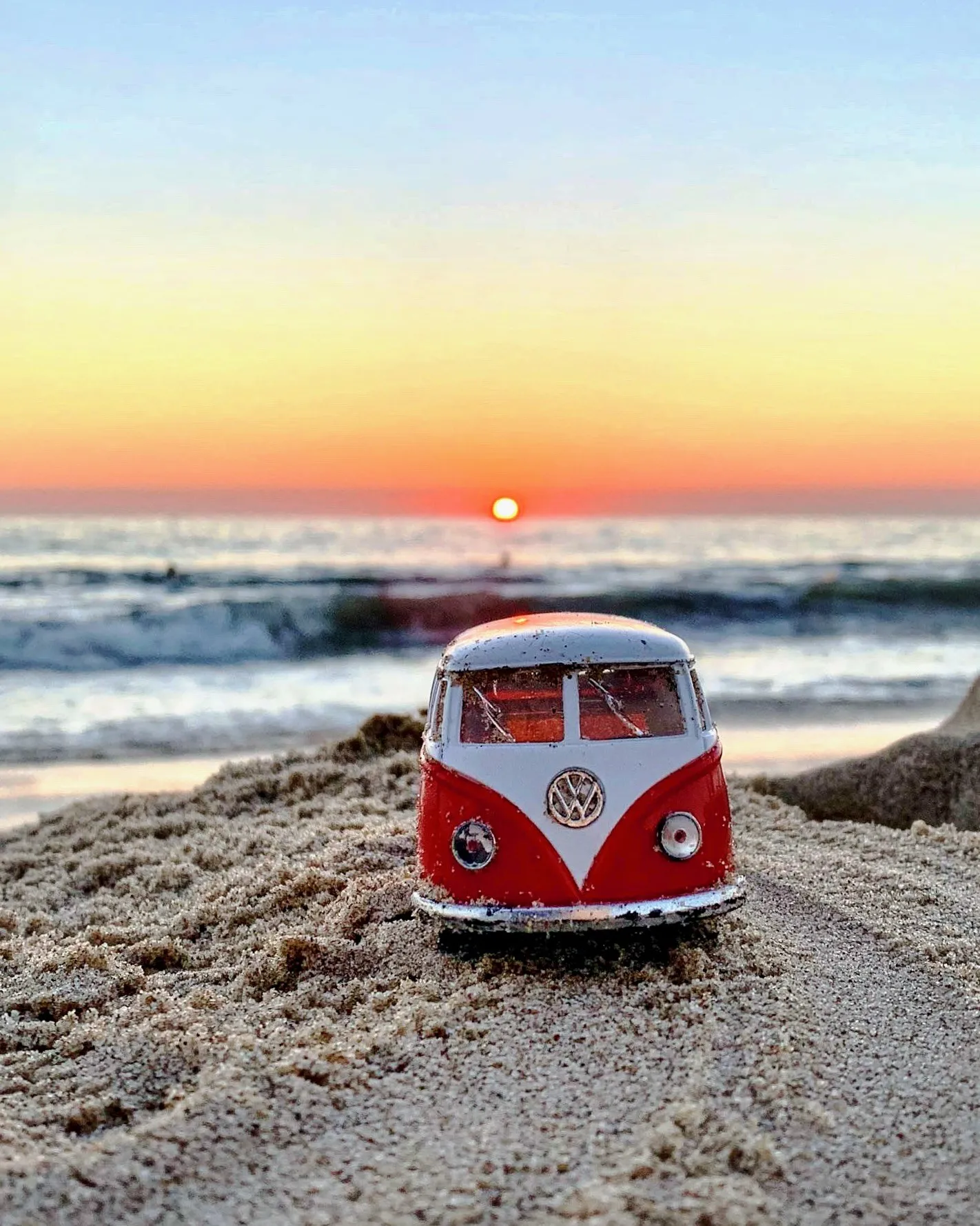 A miniature red and white Volkswagen van on the sand beneath the setting sun
