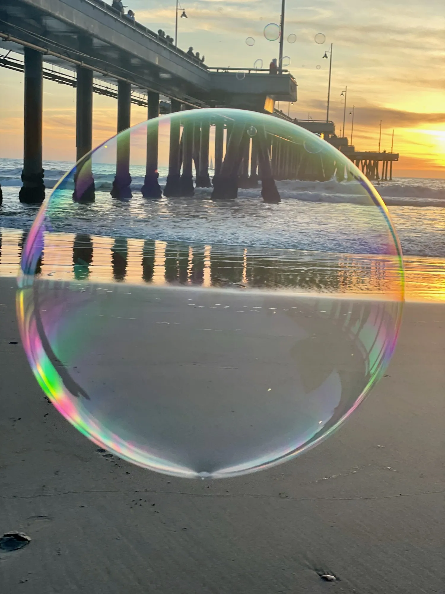 A large iridescent soap bubble framing the pier at sunset