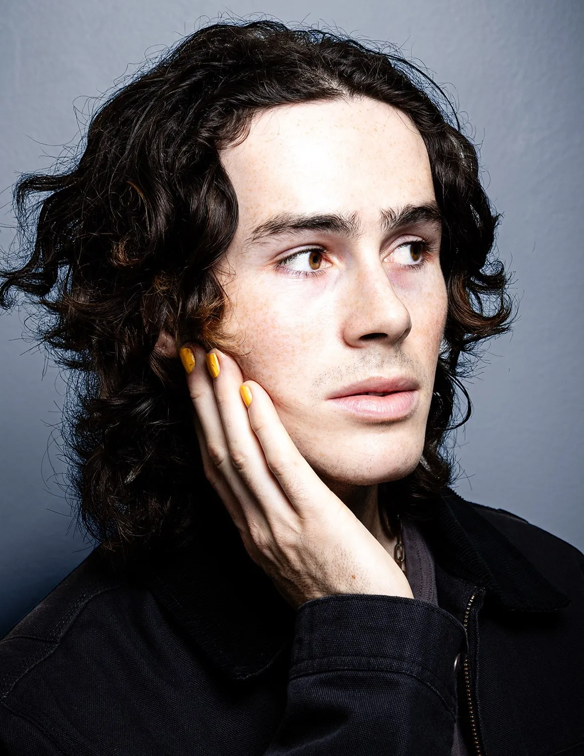A curly-haired man resting his cheek on his hand against a gray background