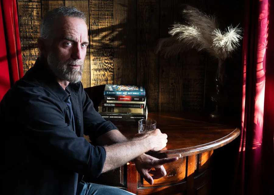 A man seated beside a bar with books and dark red curtains