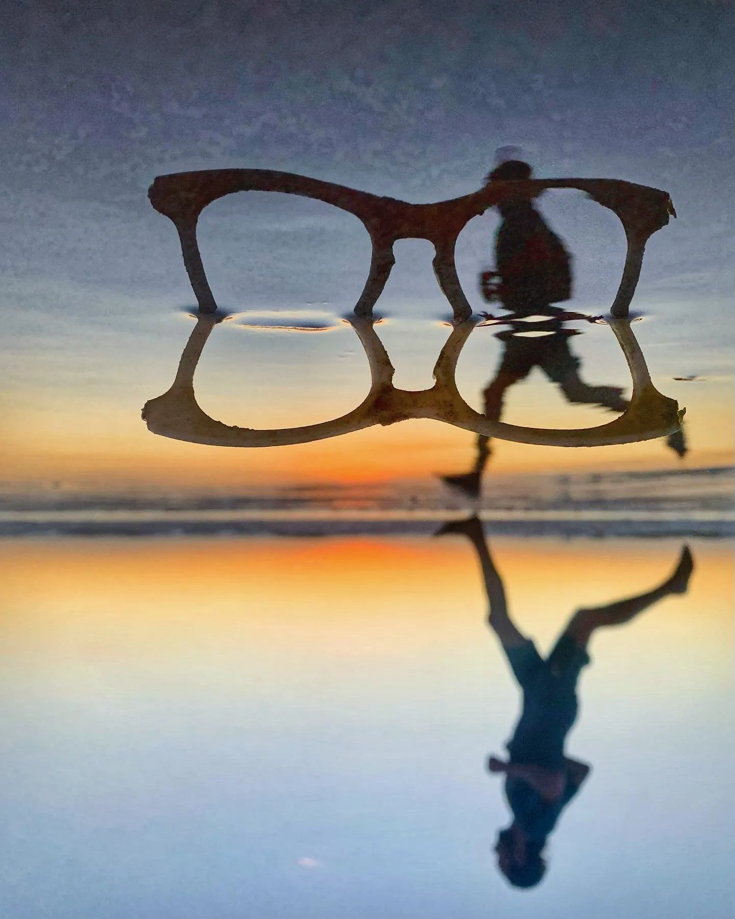 A runner appears within the reflection of oversized glasses on the beach.