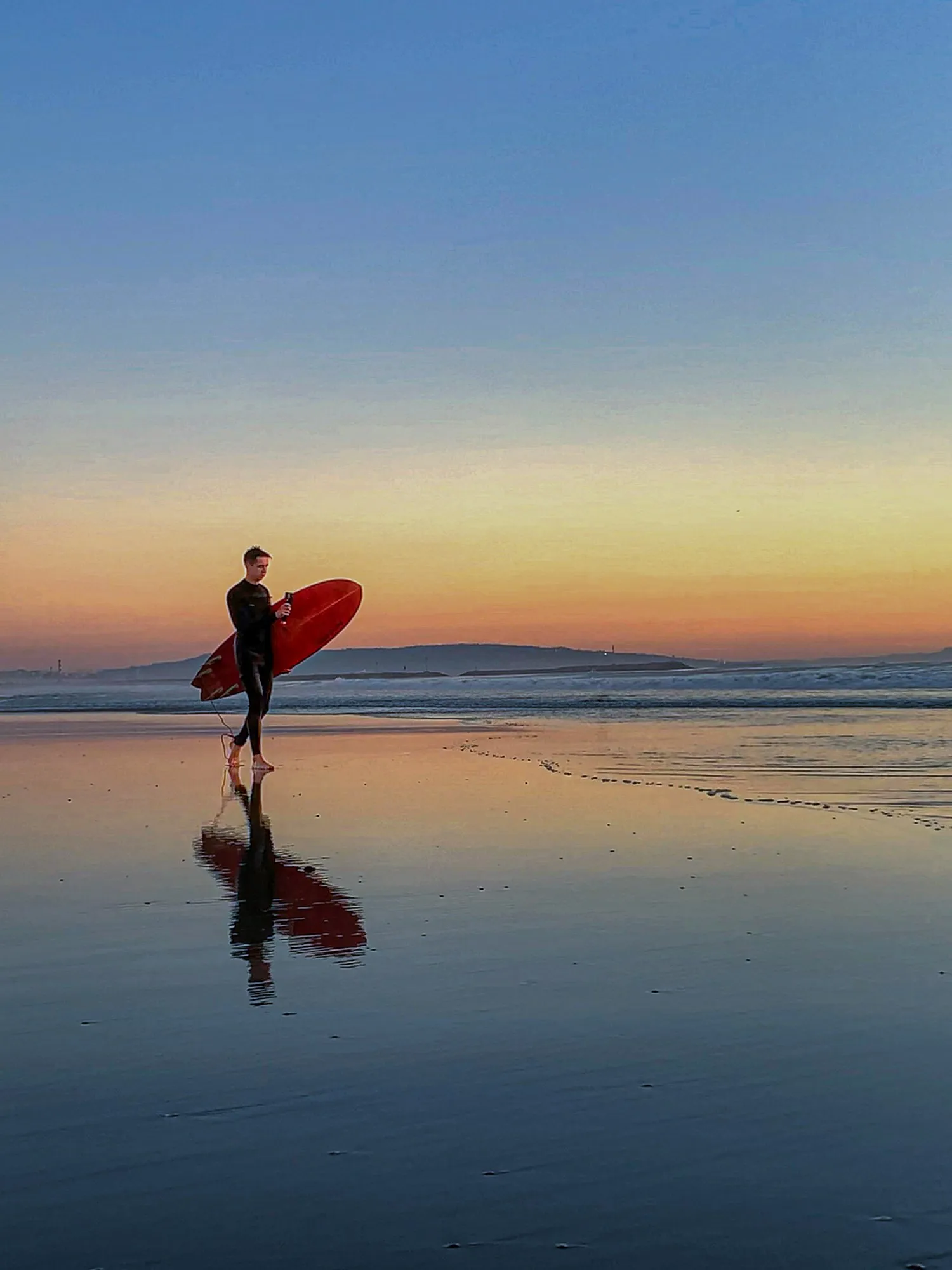 A surfer and board are reflected in wet sand beneath a violet sky.