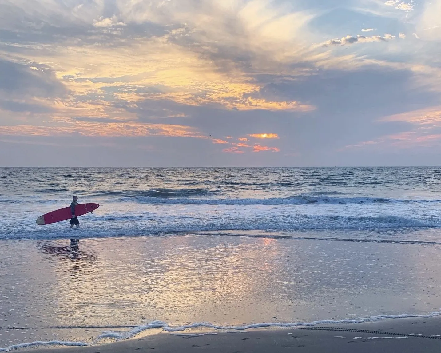 A surfer stands beside a red board as long waves meet the shore.