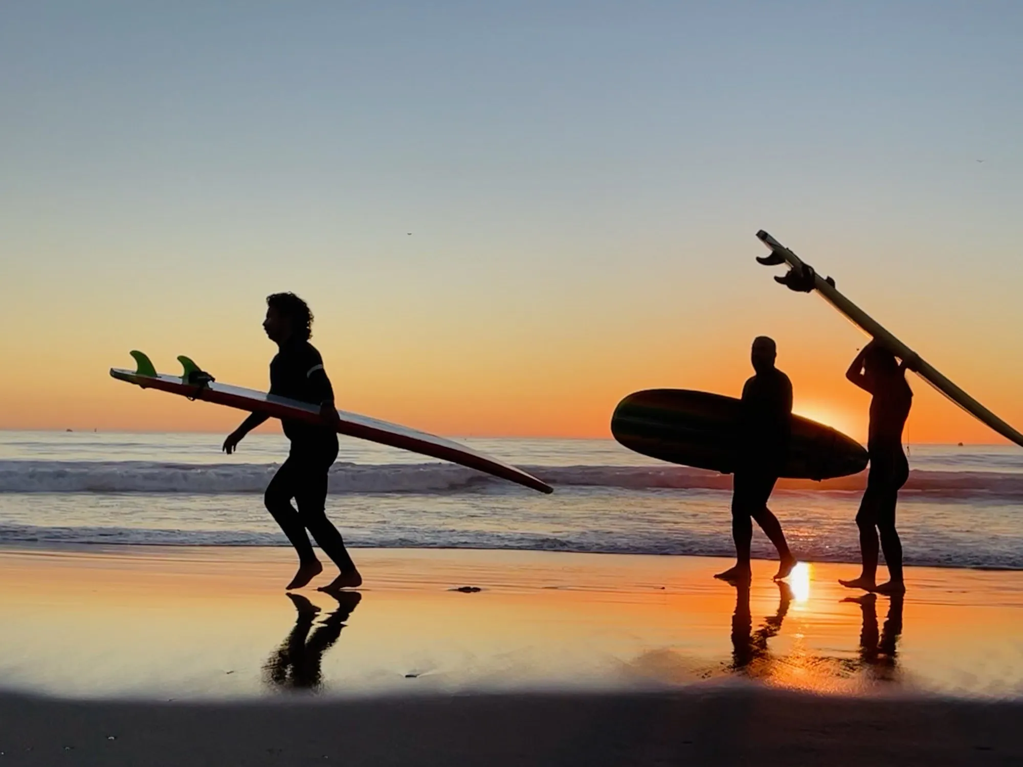Surfers carrying boards as silhouettes against an orange horizon