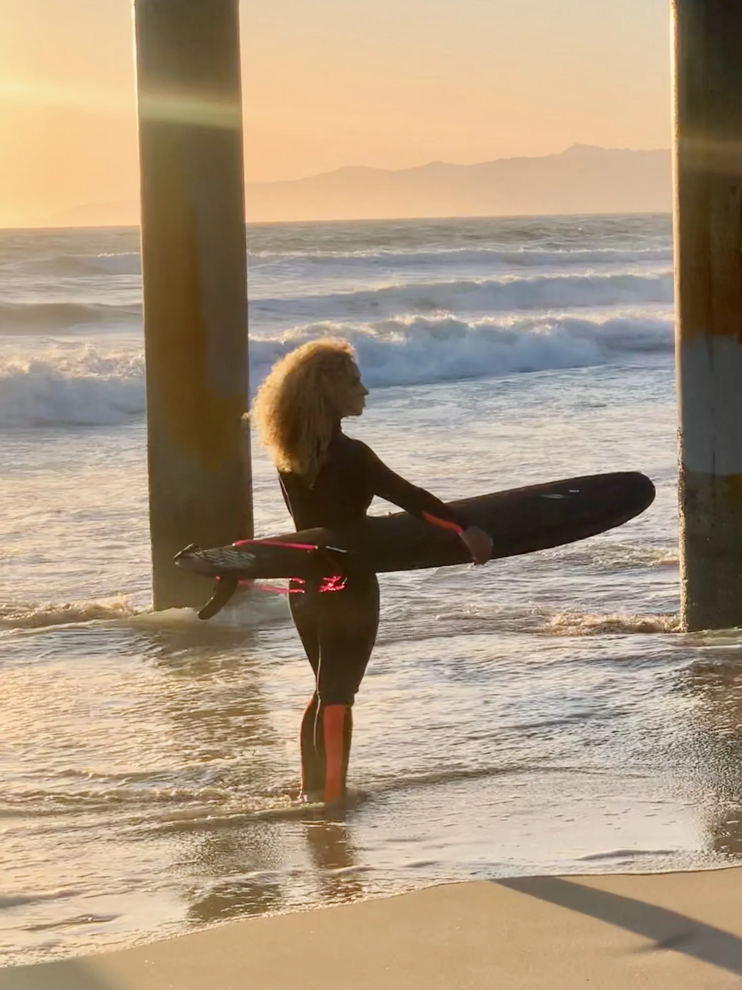 A surfer carrying a longboard through warm light beneath a pier