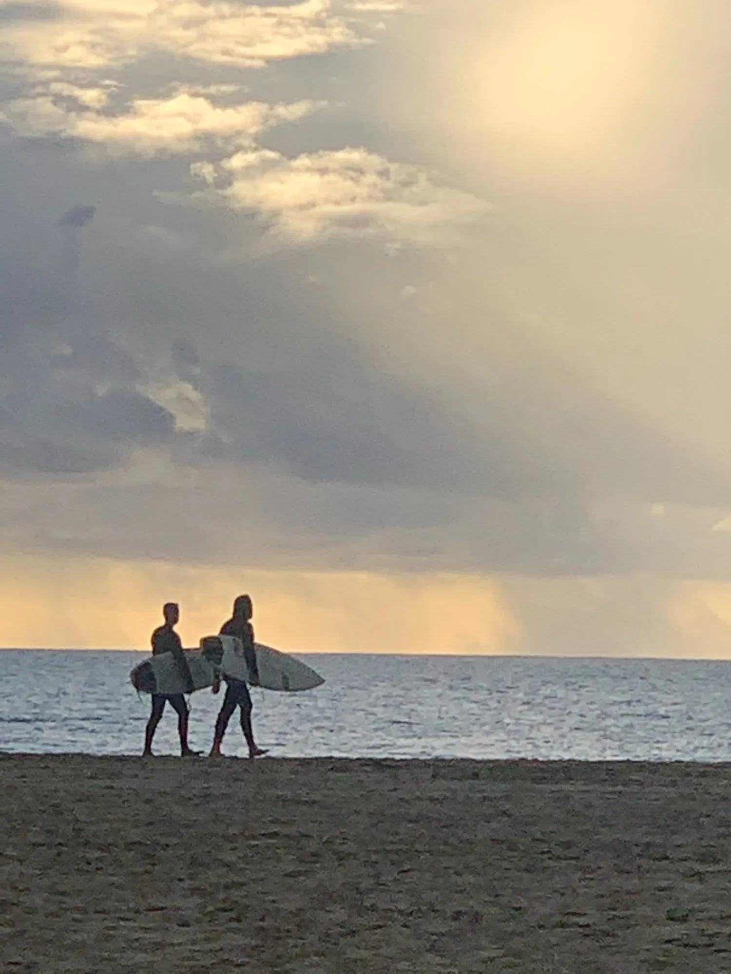 Two surfers carrying boards beneath shafts of light in a dramatic sky