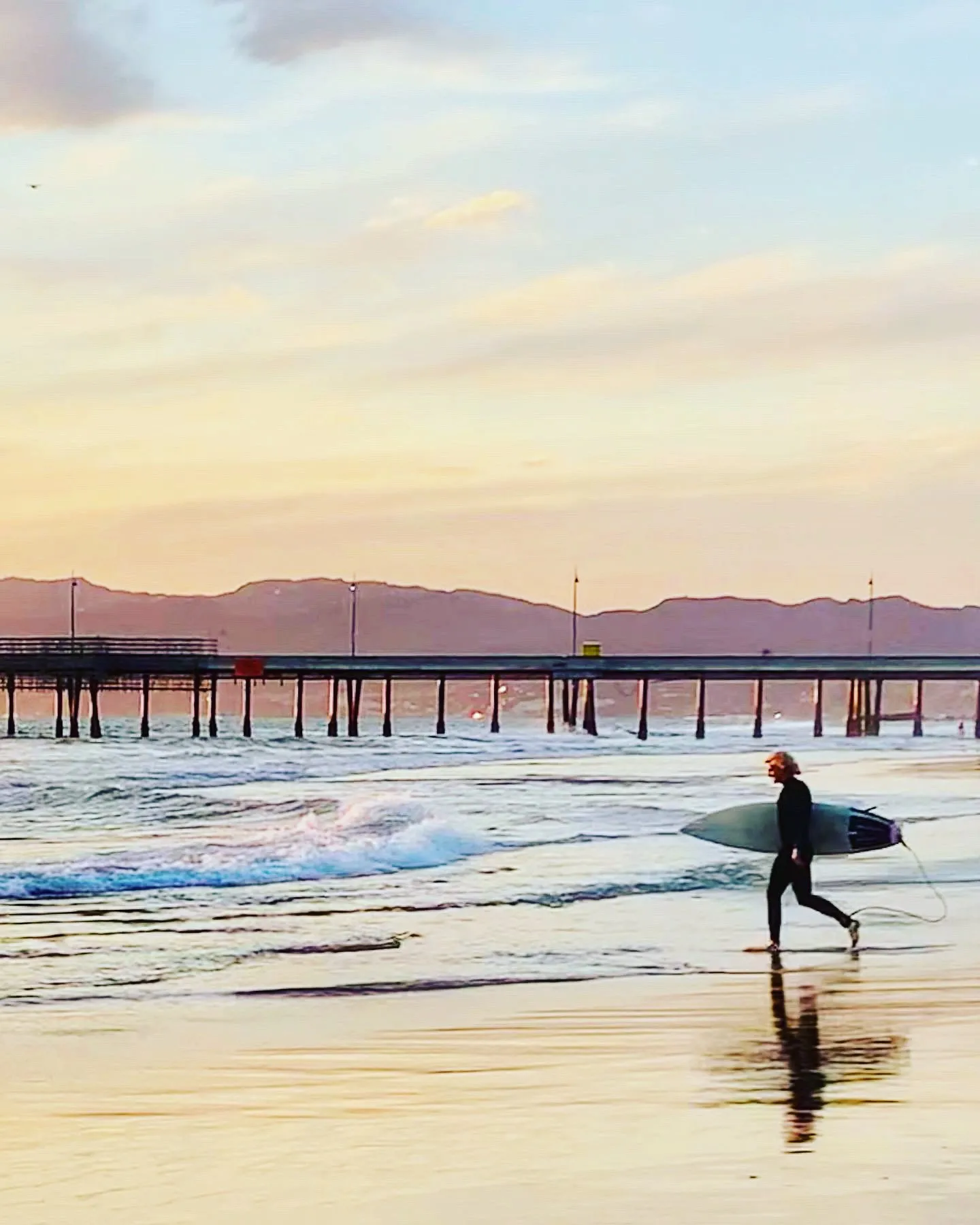 A surfer walking along reflective wet sand with a pier and mountains beyond