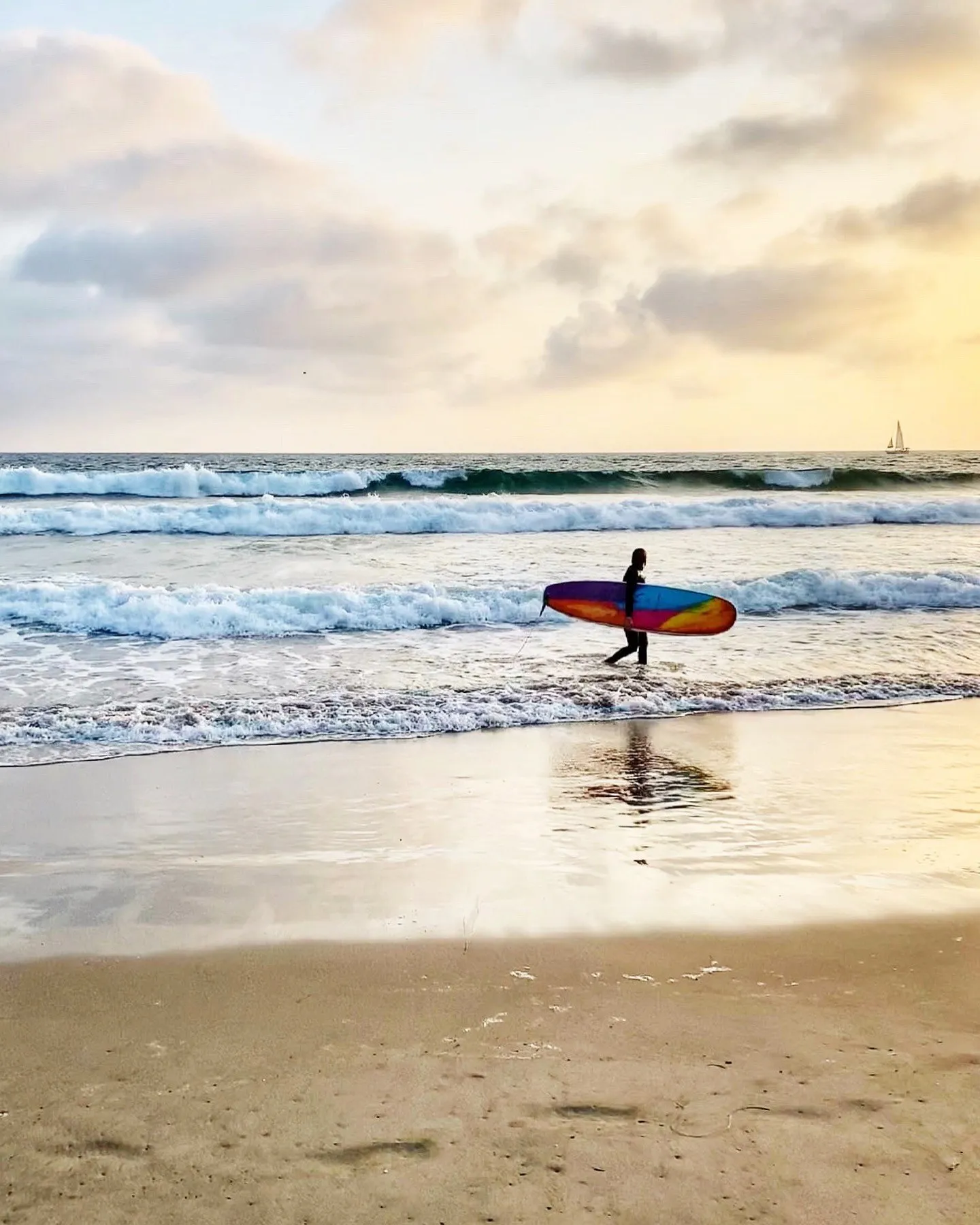 A surfer carrying a colorful board beside pale breaking waves