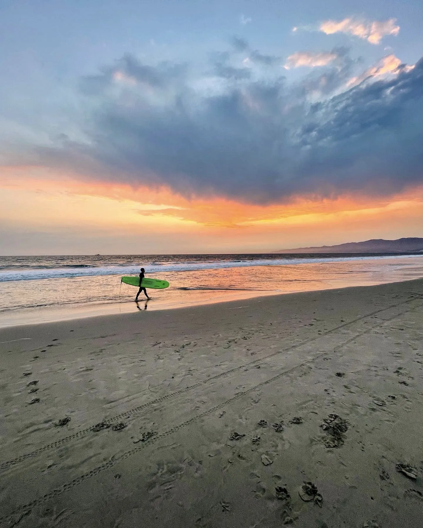 A distant surfer with a green board beneath sweeping clouds at sunset