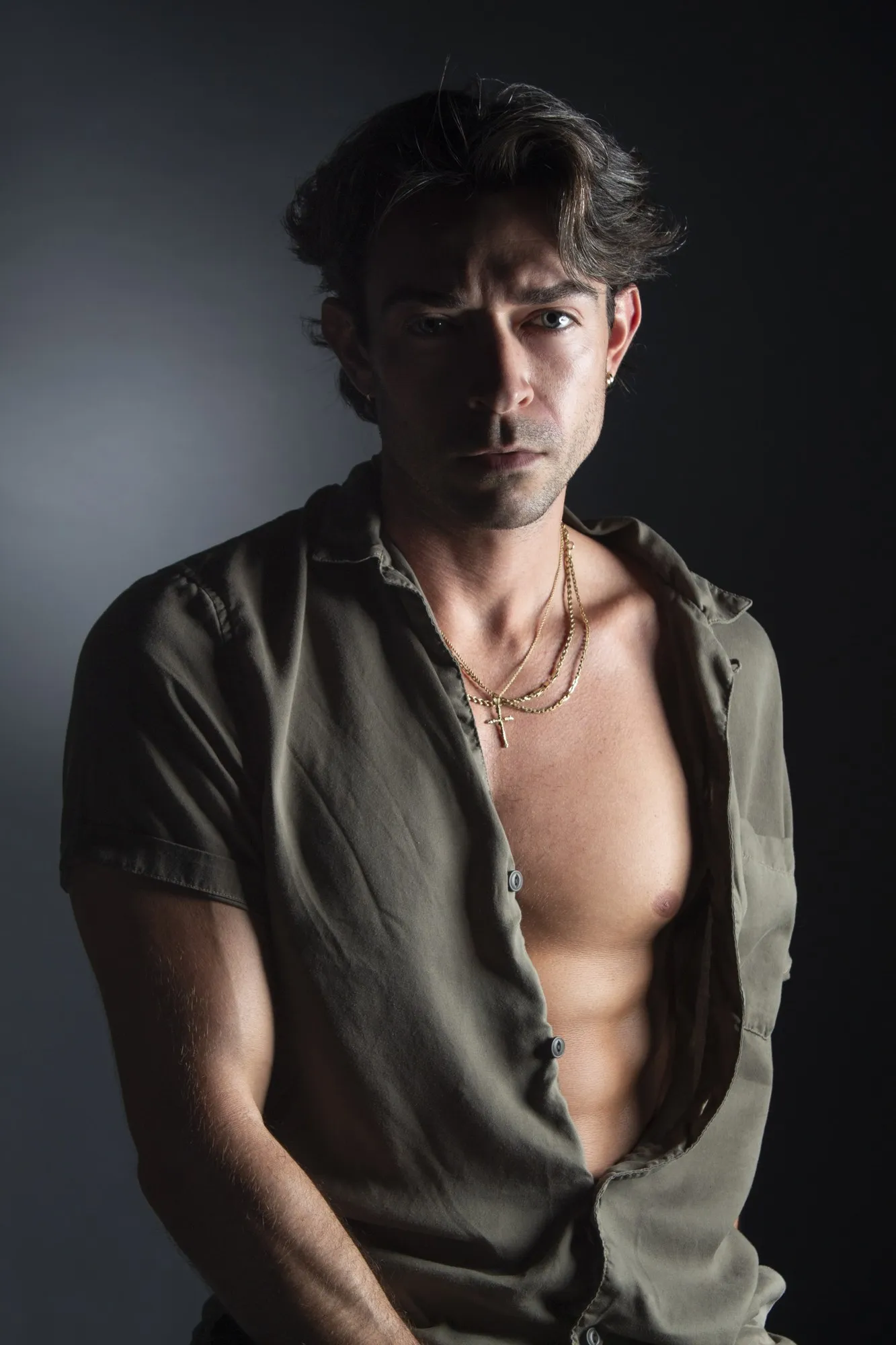 A young man in an open shirt poses against a dark studio background.
